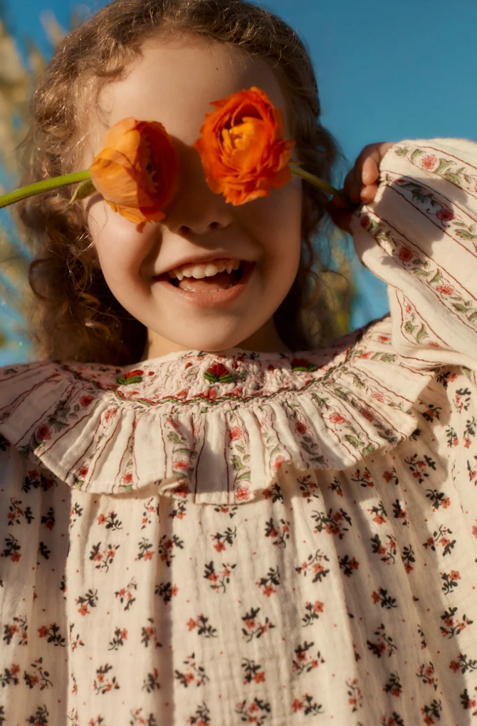 A close-up lifestyle shot of a young girl outdoors wearing a cream-colored floral dress with a ruffled collar. She is smiling and holding two orange flowers in front of her eyes, with a bright blue sky in the background.